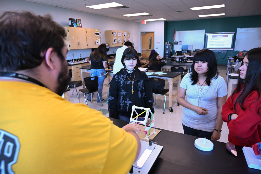 In a classroom, a man in a yellow shirt speaks with a student holding a model. Other students are seated at tables.