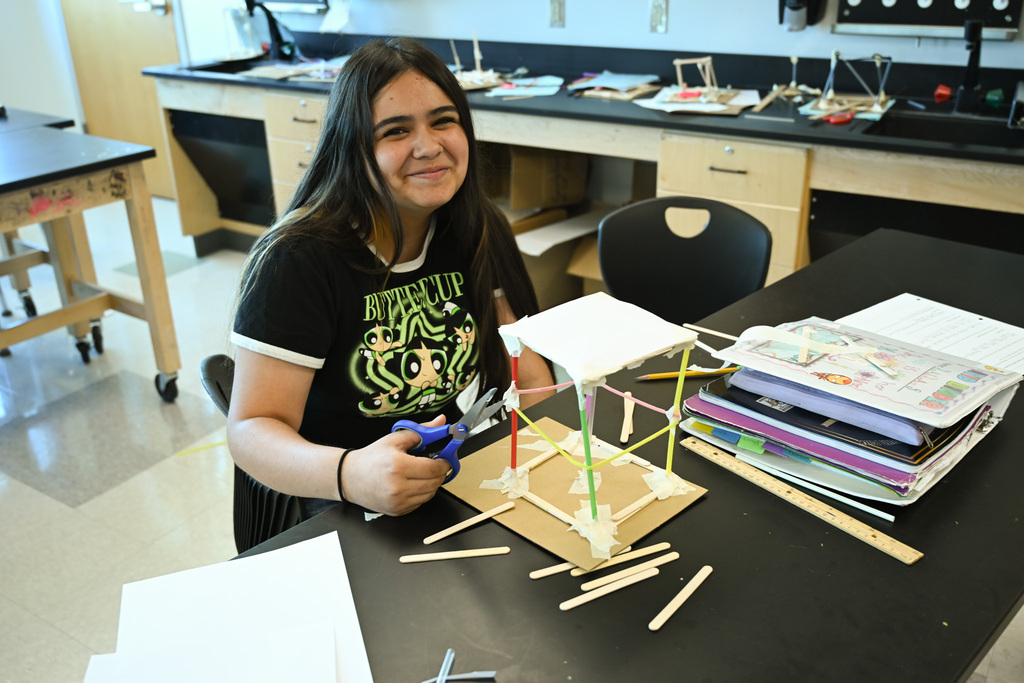 A woman in a black t-shirt works on a model structure in a classroom.