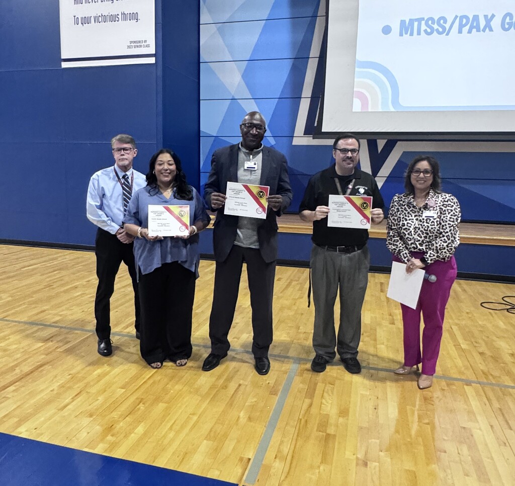 Five people stand in a row in a gym, holding certificates. Behind them is a wall with a sign and a screen.
