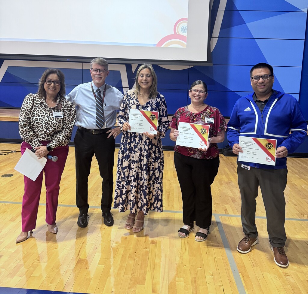 Five people stand on a basketball court, holding certificates. Three wear glasses. A screen is above them.