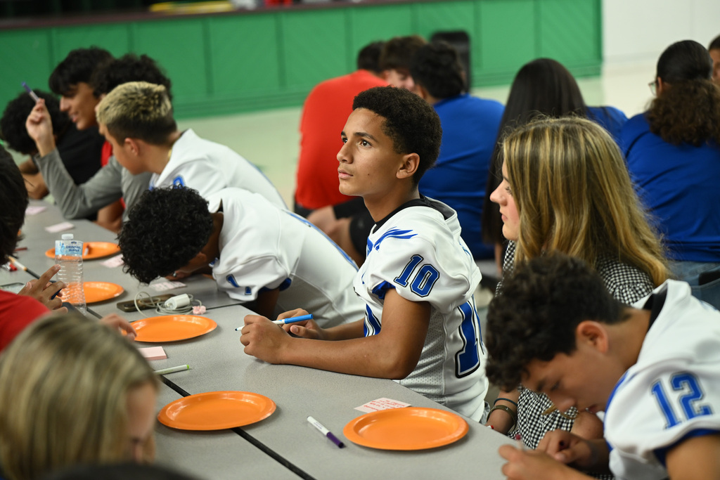 A group of young people in jerseys sit at a table. They have plates and pens.