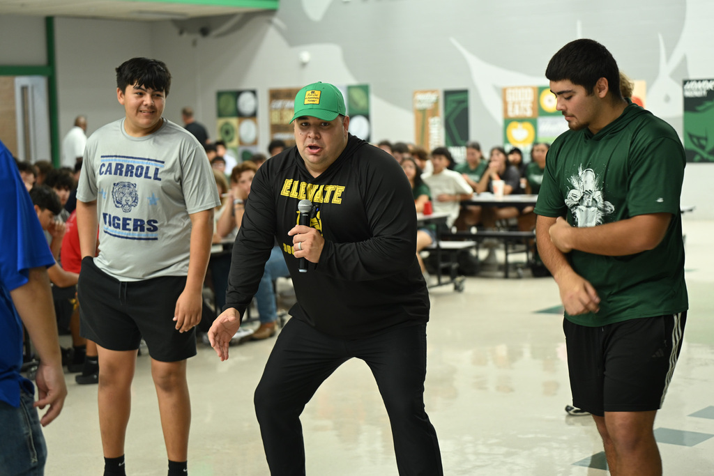 Three individuals stand in a cafeteria. The center person holds a microphone, speaking. Background includes tables, chairs, and students.