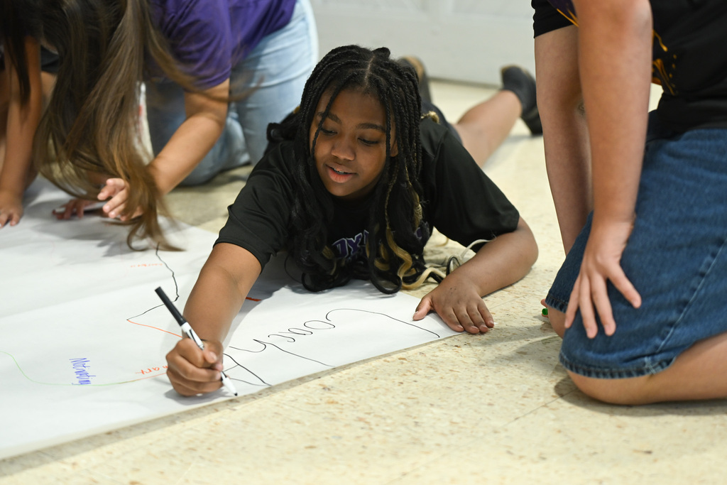 Several people on floor, one lying down drawing on paper with marker. Floor and paper visible.