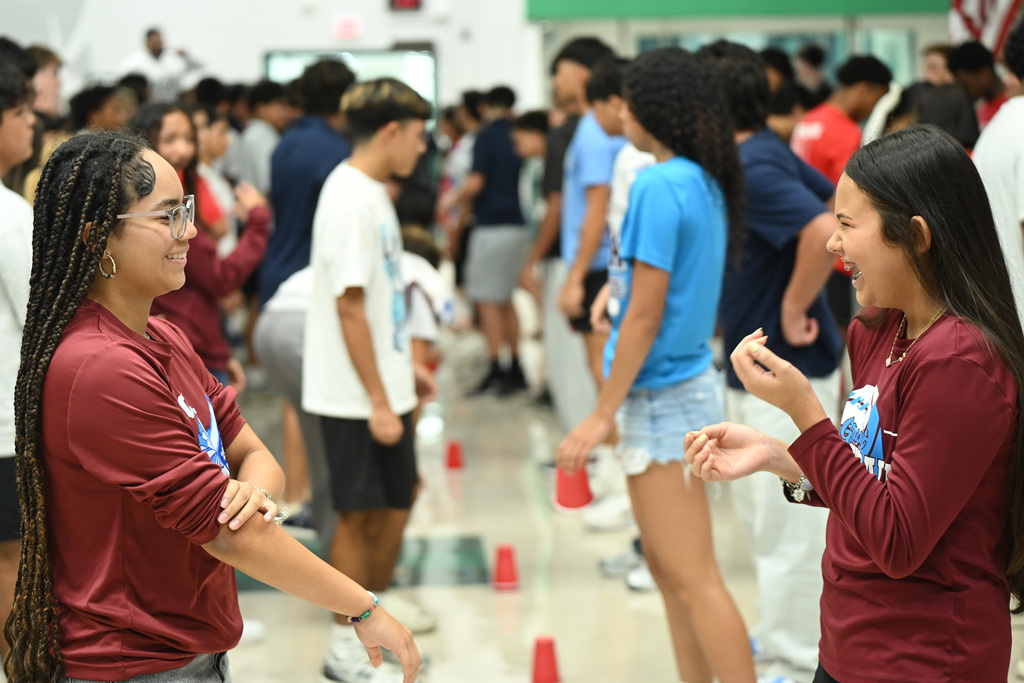 Students in maroon shirts stand and chat, facing each other, amid a crowd in a hallway.