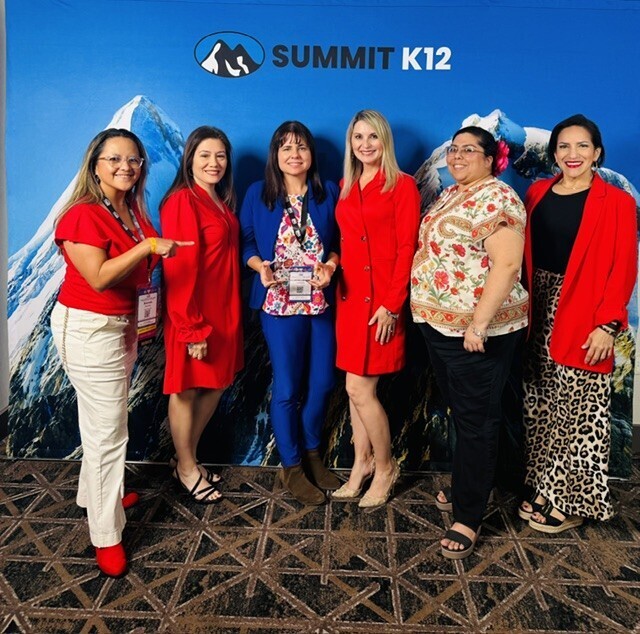 Six women pose in front of a blue backdrop with mountain design and text. They wear various colored outfits, including red, blue, and black.