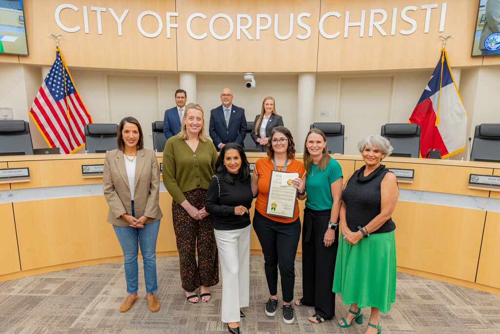 Six women, one holding a certificate, stand in a city council meeting room with flags and a podium.