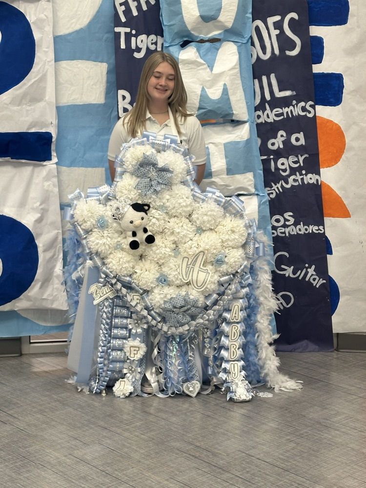 Person with blond hair, wearing white shirt, stands before blue and white backdrop. Muffler made of white and blue flowers, and black and white stuffed animal.