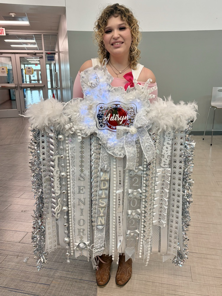 Person holding a large sash with text and ribbons in front of a hallway.