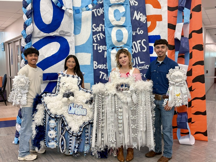Four students stand before a banner with blue, white, and orange letters, holding pom-poms.