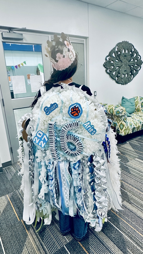 A student wears a blue and white pom-pom with a crown in a hallway next to a couch.