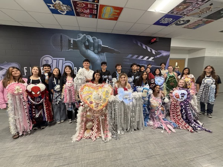 Group of students posing for photo, some wearing colorful costumes, in front of large backdrop.