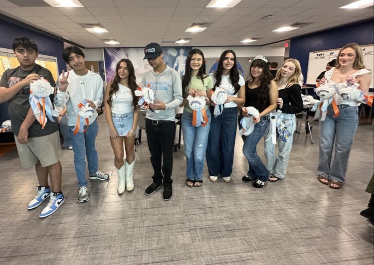 Group of students posing for a photo, holding up stuffed animals, with blue walls and tiled floor.