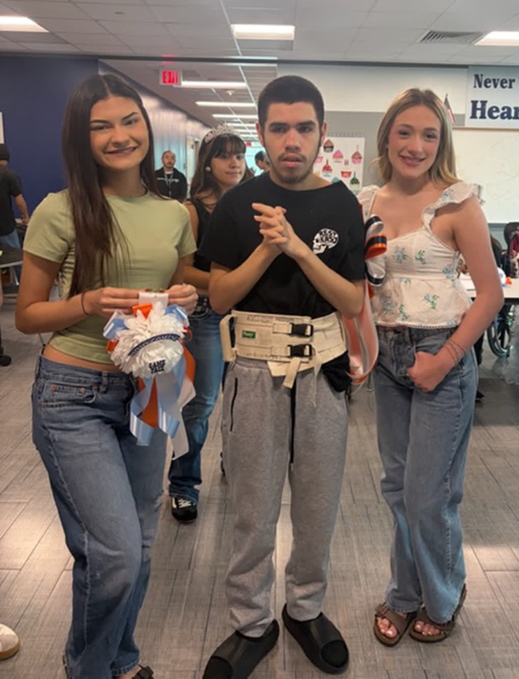 Three students standing together, one holding a white object, inside a room with tables and people walking.