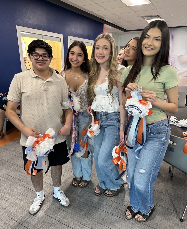 Five young people, two females and three males, stand close together in a room. They hold orange and white decorations.