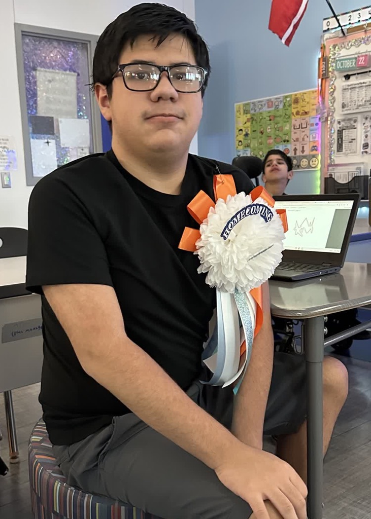 Man in black shirt sits at a desk in classroom; wearing glasses and a flower corsage.