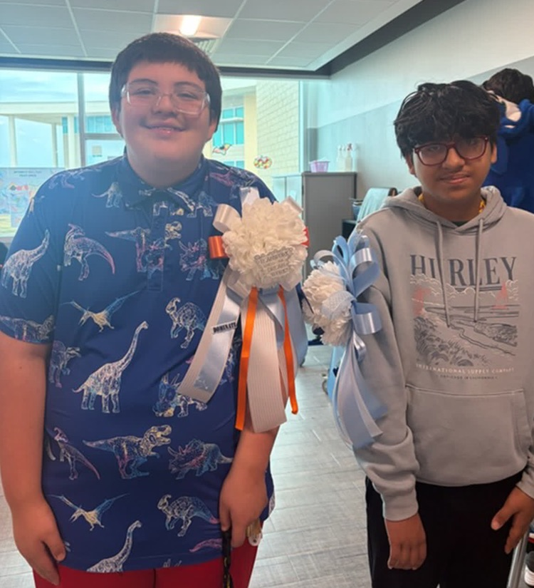 Two boys with school IDs, one holding a corsage, stand in a hallway, smiling for the camera.
