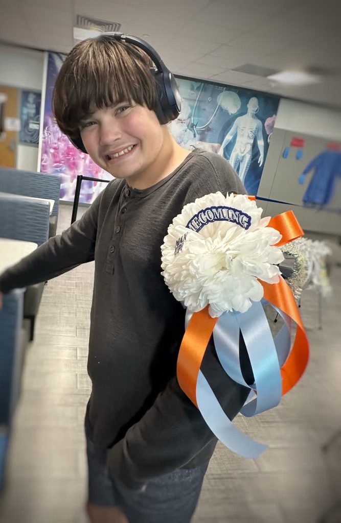 A smiling young boy wearing headphones holds a white flower with orange and blue ribbons.
