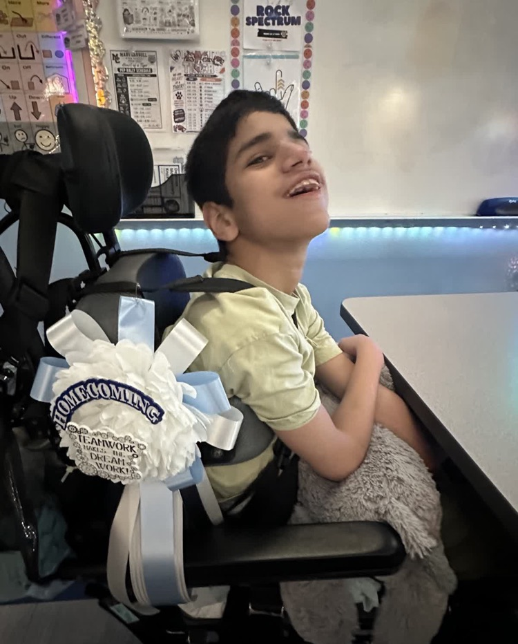 Boy in wheelchair wearing bow with words "Homecoming" sits smiling in classroom. White wall behind.