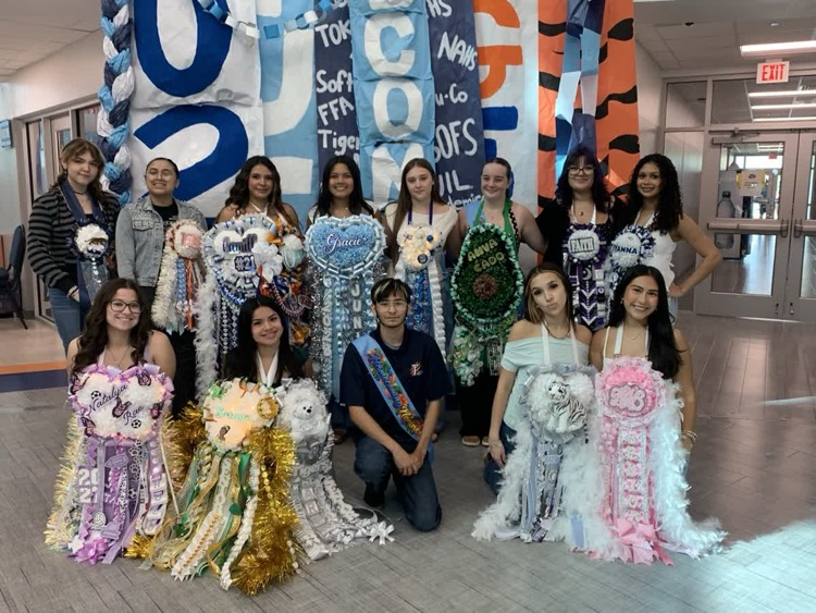 A group of young people in colorful costumes, some with sashes, pose in front of a decorated wall.
