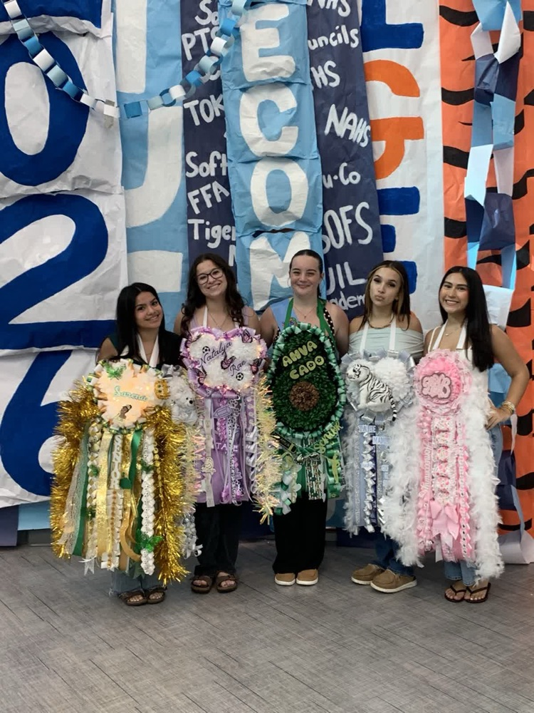 Five young women in costumes stand side by side, holding handmade items, in front of a decorated wall.