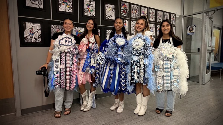 Five young women in matching outfits stand together in a room with artwork on the wall.