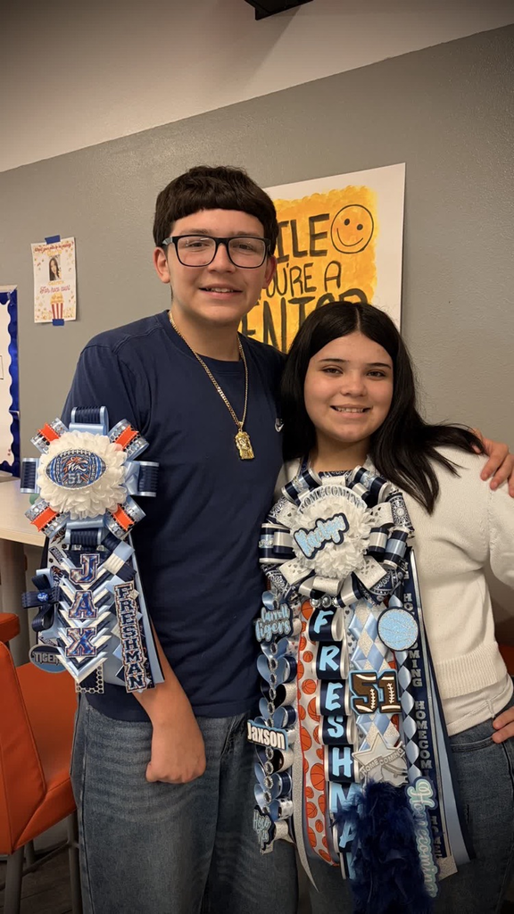 Two students stand in a room. A boy wears glasses and a blue shirt. A girl has a white sweater and holds a sash with ribbons and words. Behind them, a wall has posters.