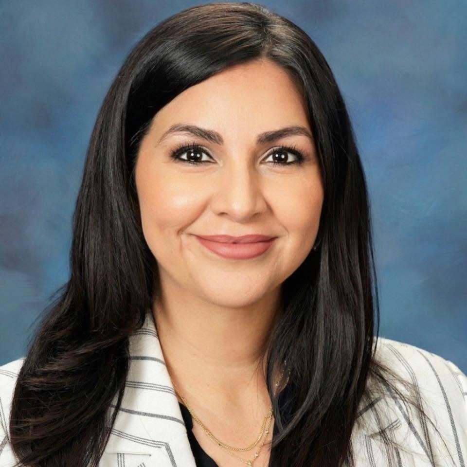 Portrait of a woman with long black hair smiling, wearing a white striped jacket, against a blue background.