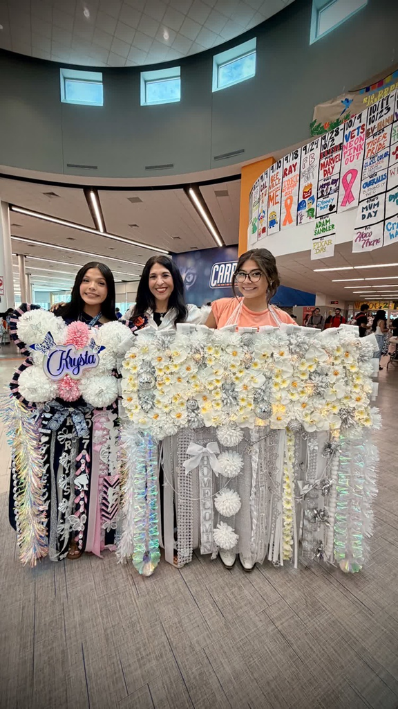 Three women pose behind a decorative banner of flowers and streamers, one wearing a sash, in a large hallway with many signs on the wall.