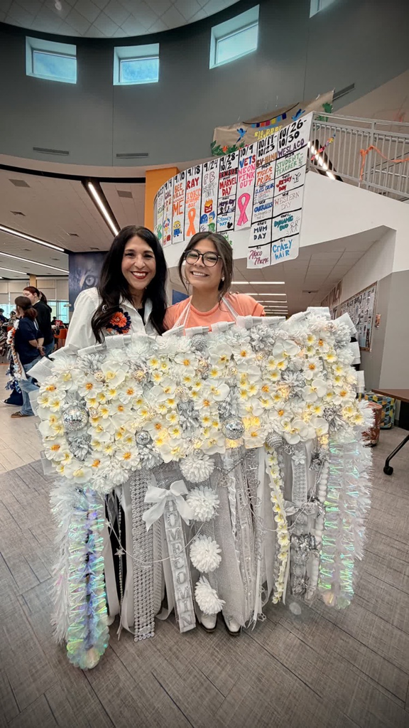 Two women stand next to each other holding a large white and yellow floral banner. Behind them, a staircase is visible.