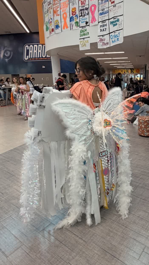 A student wearing glasses walks through a mall, sporting wings, a butterfly costume, and a white cape.
