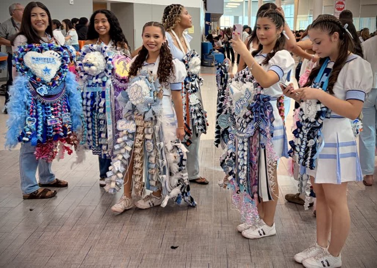 Students wearing blue and white cheer uniforms holding blue and white pom-poms in a hallway.