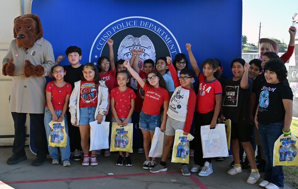 Group of children holding bags, standing in front of a blue banner with a logo.