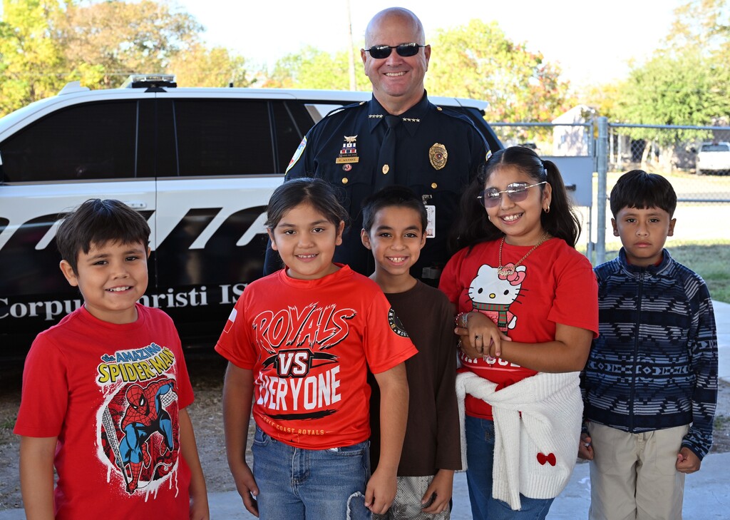 Five young children and an adult stand next to a police vehicle. The adult wears sunglasses and a police uniform.