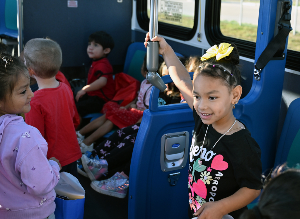 Children sit inside a blue bus with blue seats; one girl holds the pole and smiles.