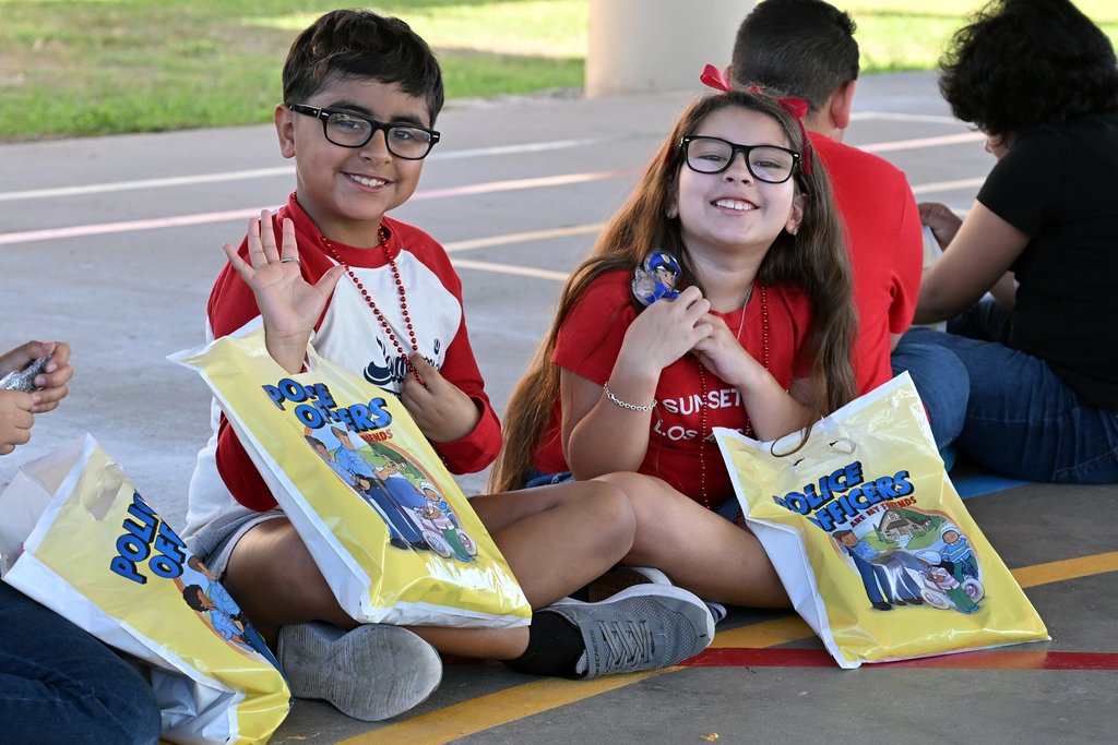 Three kids and an adult sit on the ground. Each child holds a yellow bag with cartoon characters. One kid has glasses and smiles.