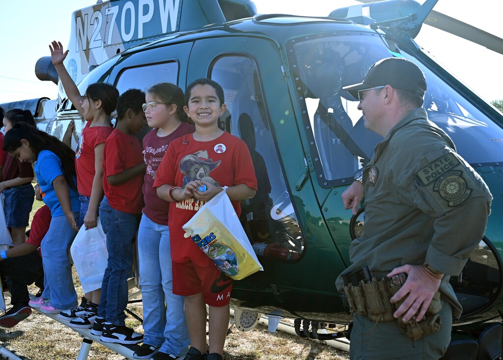 Several students wearing red shirts stand near a helicopter with a person in a green uniform and hat.