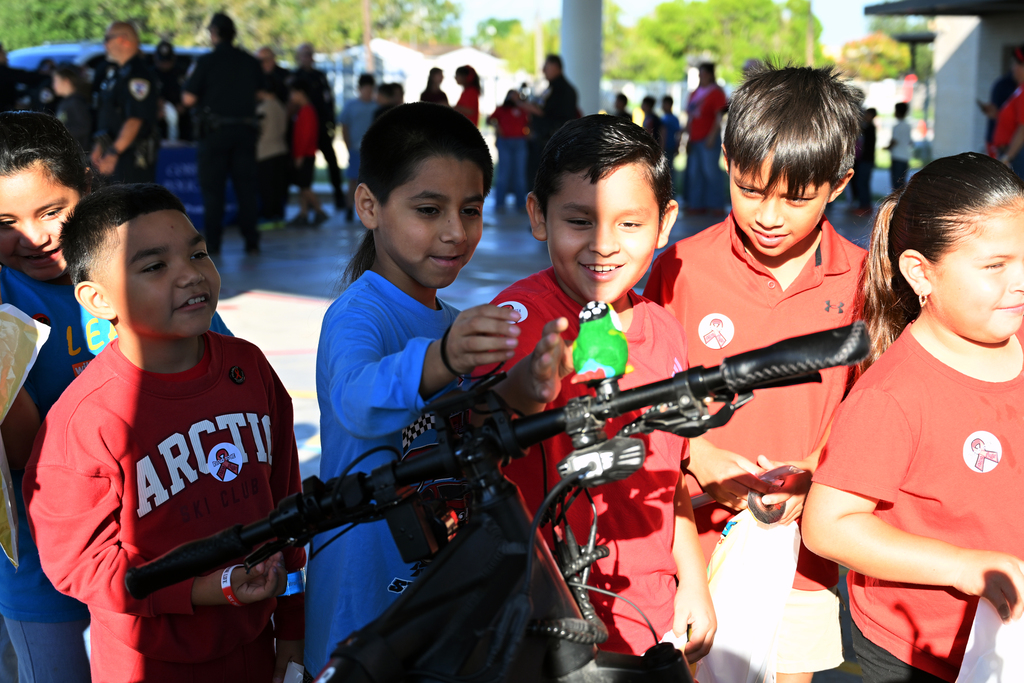 Children wearing red shirts gather around a bike. One holds a green bottle. A large group stands behind.