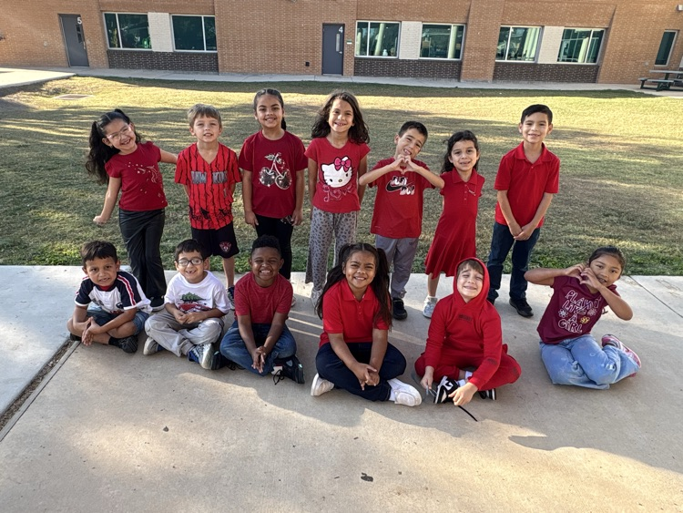 A group of children in red shirts, seated and standing, posing for a photo in front of a building.