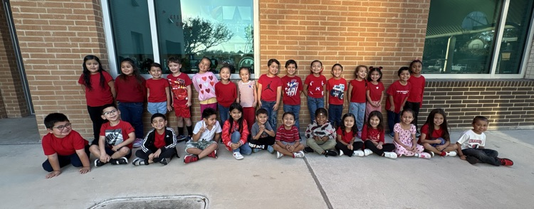 Group of children in red shirts sitting and kneeling in front of a brick building.