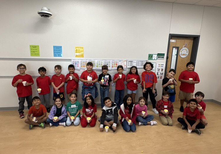 A classroom with wood floors, children seated, each holding a white cup, all wearing red shirts, a closed door in the background.