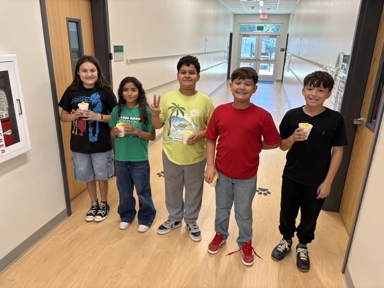 Five children stand in a hallway, holding ice cream cups. They wear casual clothing and sneakers.