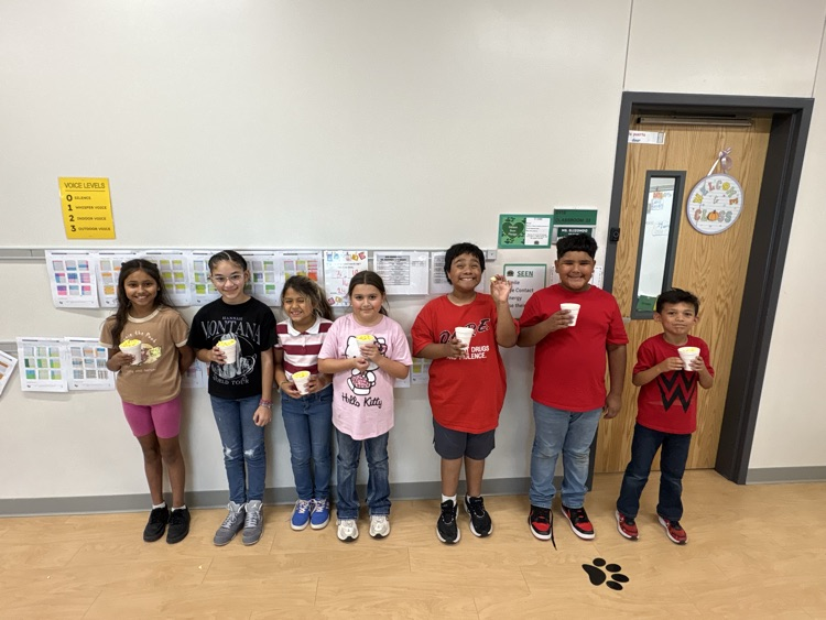 Seven children standing in a row in a classroom, each holding a cup with food in it.