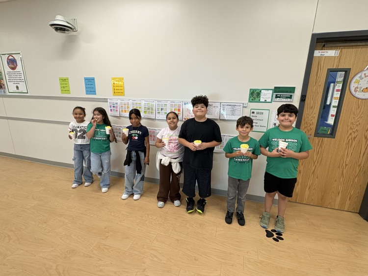Students standing in a line, holding cups, with a teacher in front, next to a door.