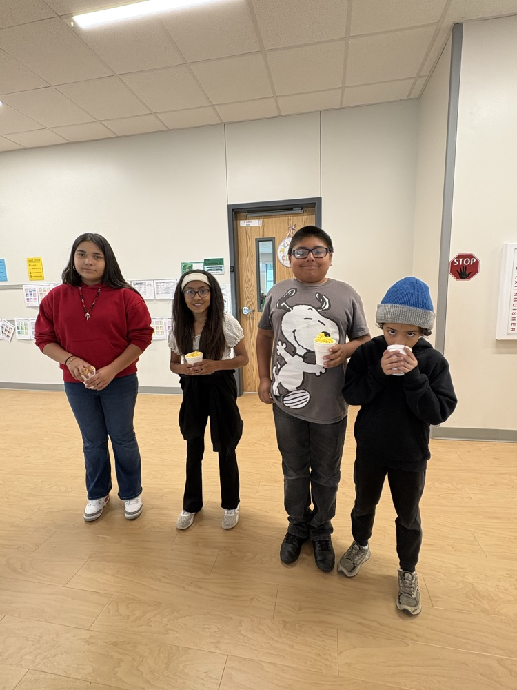 Four children stand in a room, each holding a cup. The room has white walls and a wooden floor.