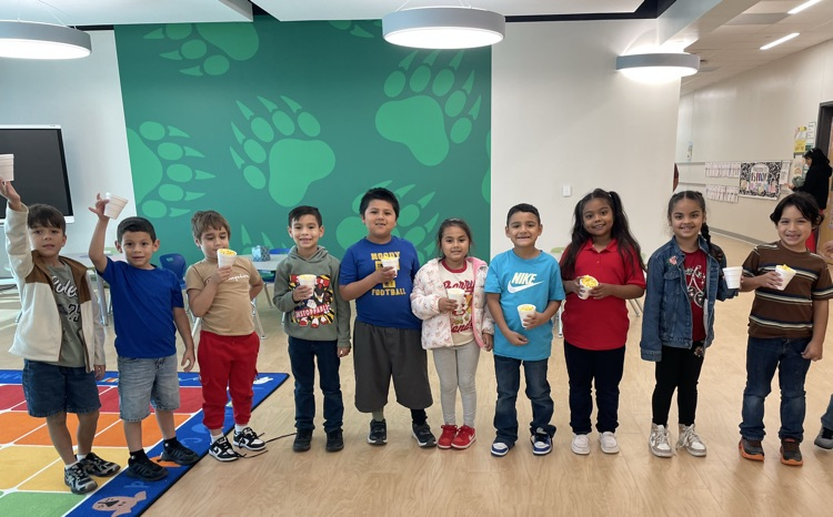 Kids stand in a line with their hands raised, each holding a cup. They stand on a floor with a colorful rug and a wall with paw prints.