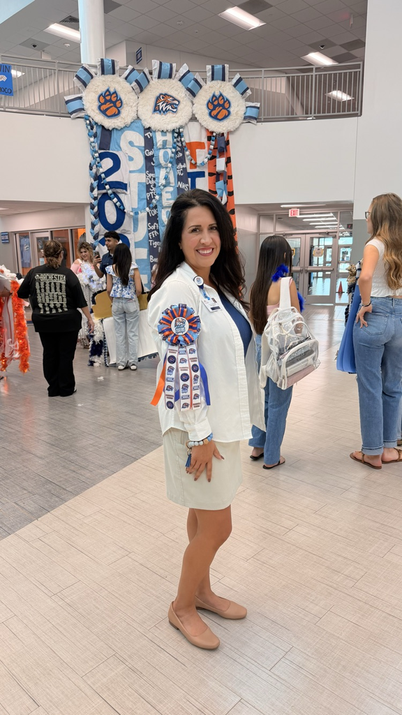 Woman stands in hallway with decorative banner behind her, smiling and looking at camera. People walk in background, two carrying backpacks.