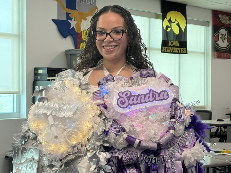A smiling woman stands, wearing glasses and holding a large, decorative, purple and silver corsage reading "Sandra".