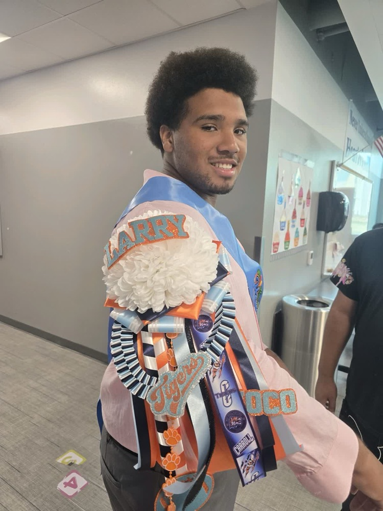 Person in school hallway, wearing sash with ribbons and flowers. Another person is in the background.