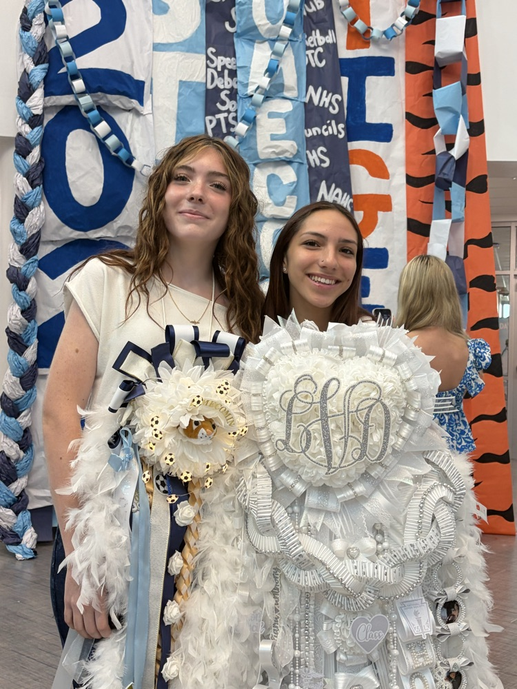 Two girls stand in front of a school banner, one holds a large white and blue sash.