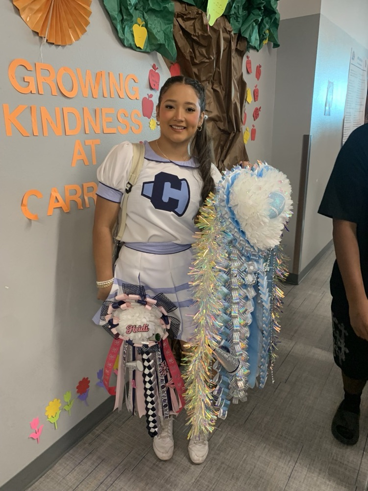 A young woman in a cheerleading uniform stands in a hallway next to a decorated wall.